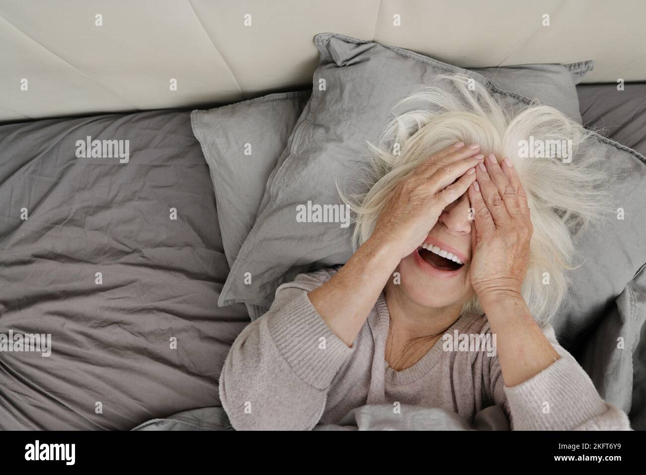 From above of anonymous elderly lady with white hair in sweater lying on comfortable soft bed and covering face with arms Stock Photo