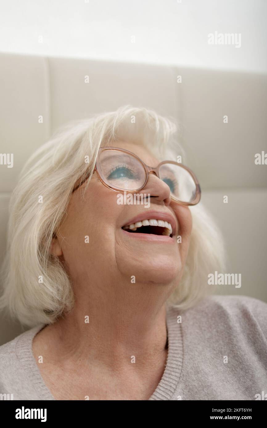Happy senior woman in eyeglasses and sweater sitting on comfortable soft bed Stock Photo