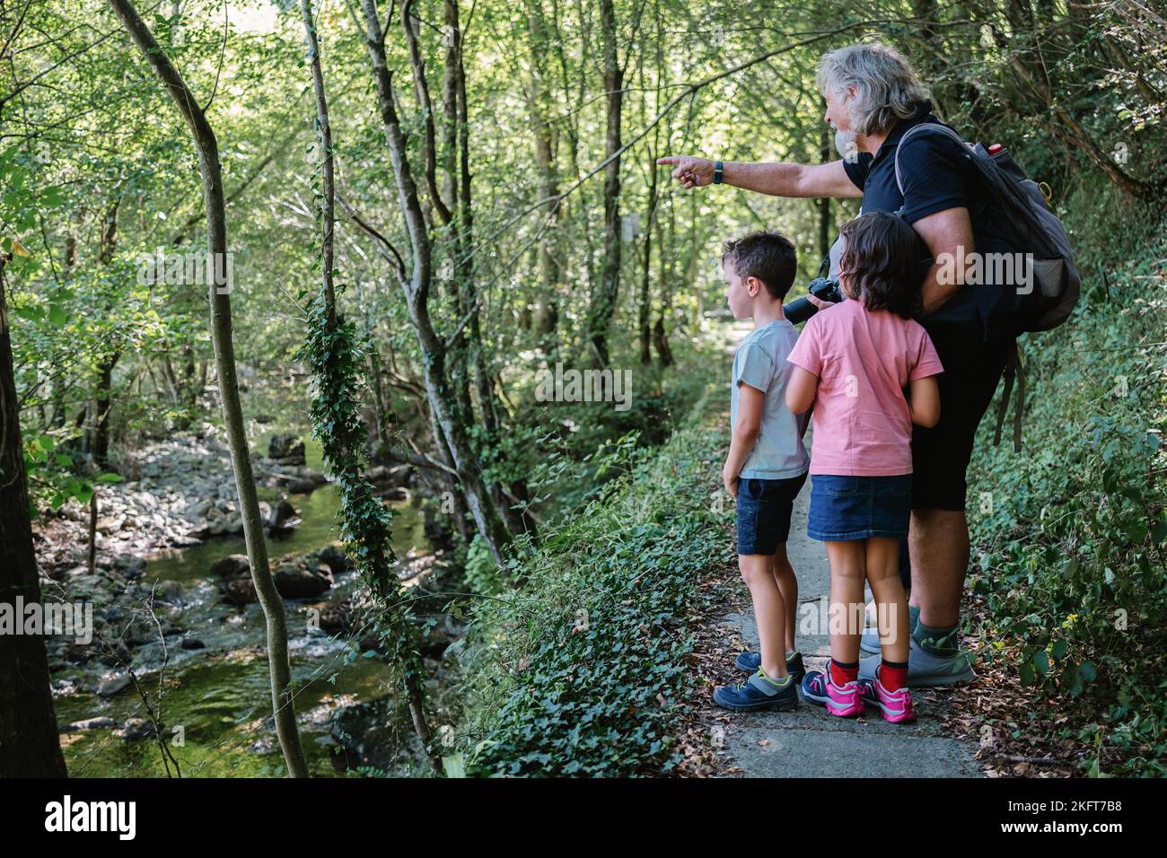 Side view full body of aged man with backpack and photo camera strolling along green forest with kids Stock Photo