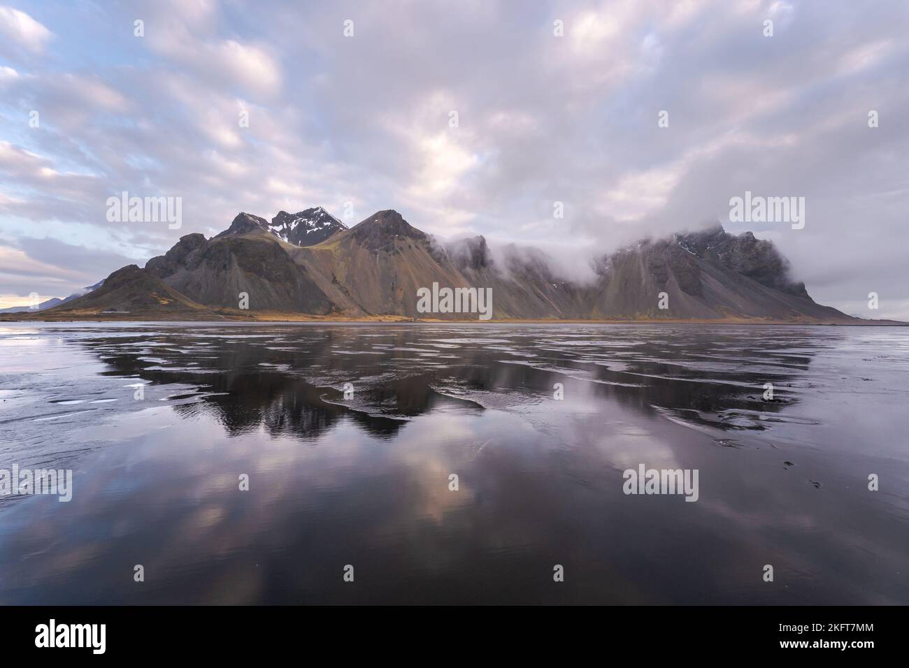 Spectacular Nordic scenery of calm frozen lake near rocky Vestrahorn Mountain with snowy peaks against cloudy sky at Stockness beach, Iceland Stock Photo