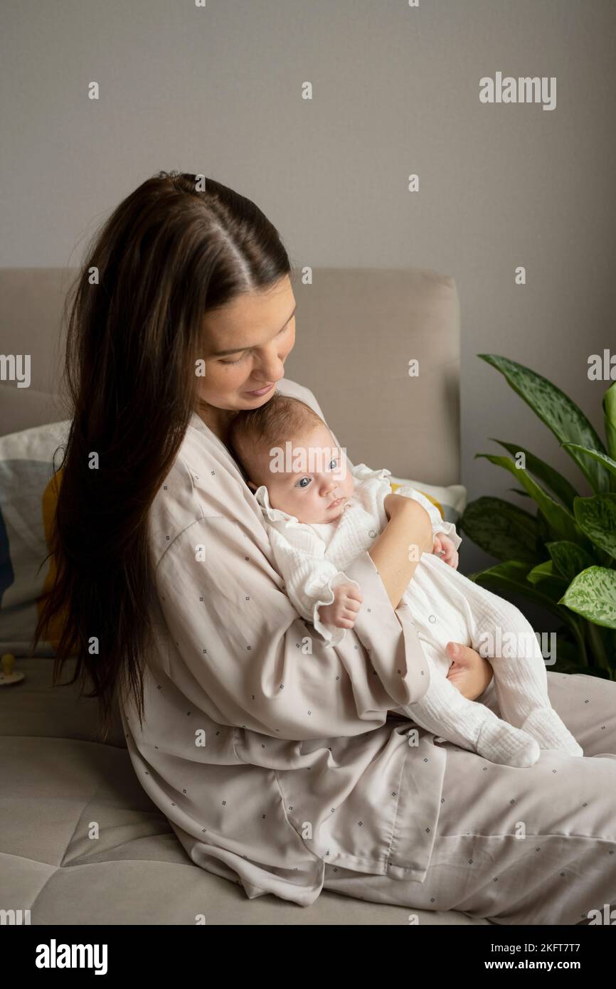 Side view of young mother in pajama holding newborn baby while sitting on sofa in morning at home Stock Photo