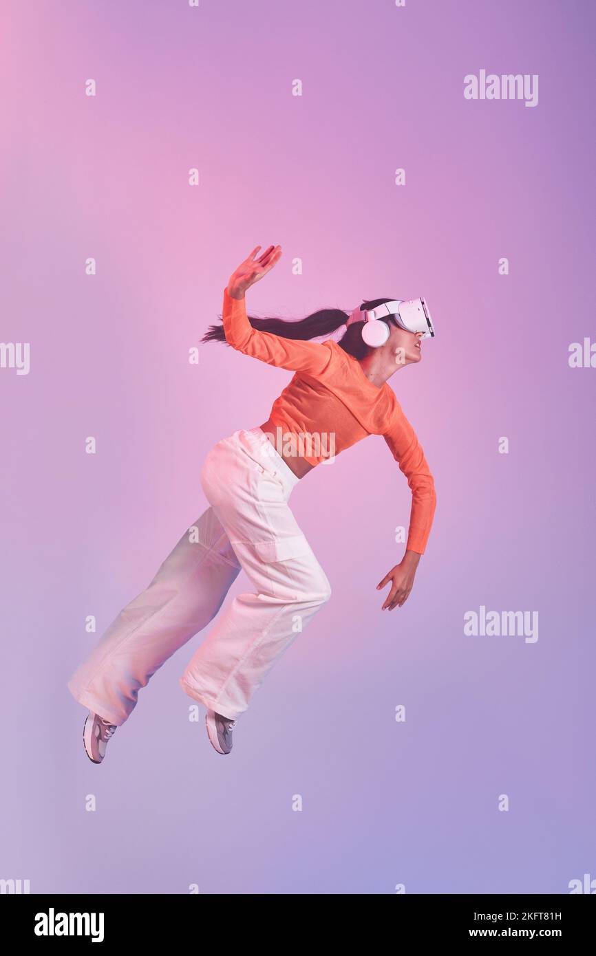 Full body female in VR helmet gesticulating and falling down against violet background while exploring cyberspace Stock Photo