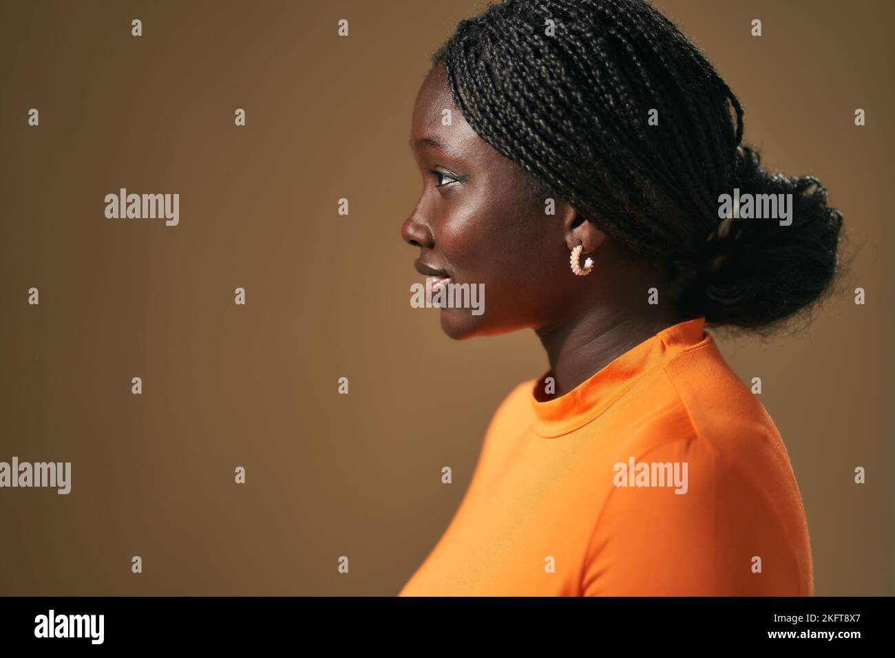 Side view portrait of smiling young African American female with Afro braids in orange top standing looking away on brown background in studio Stock Photo