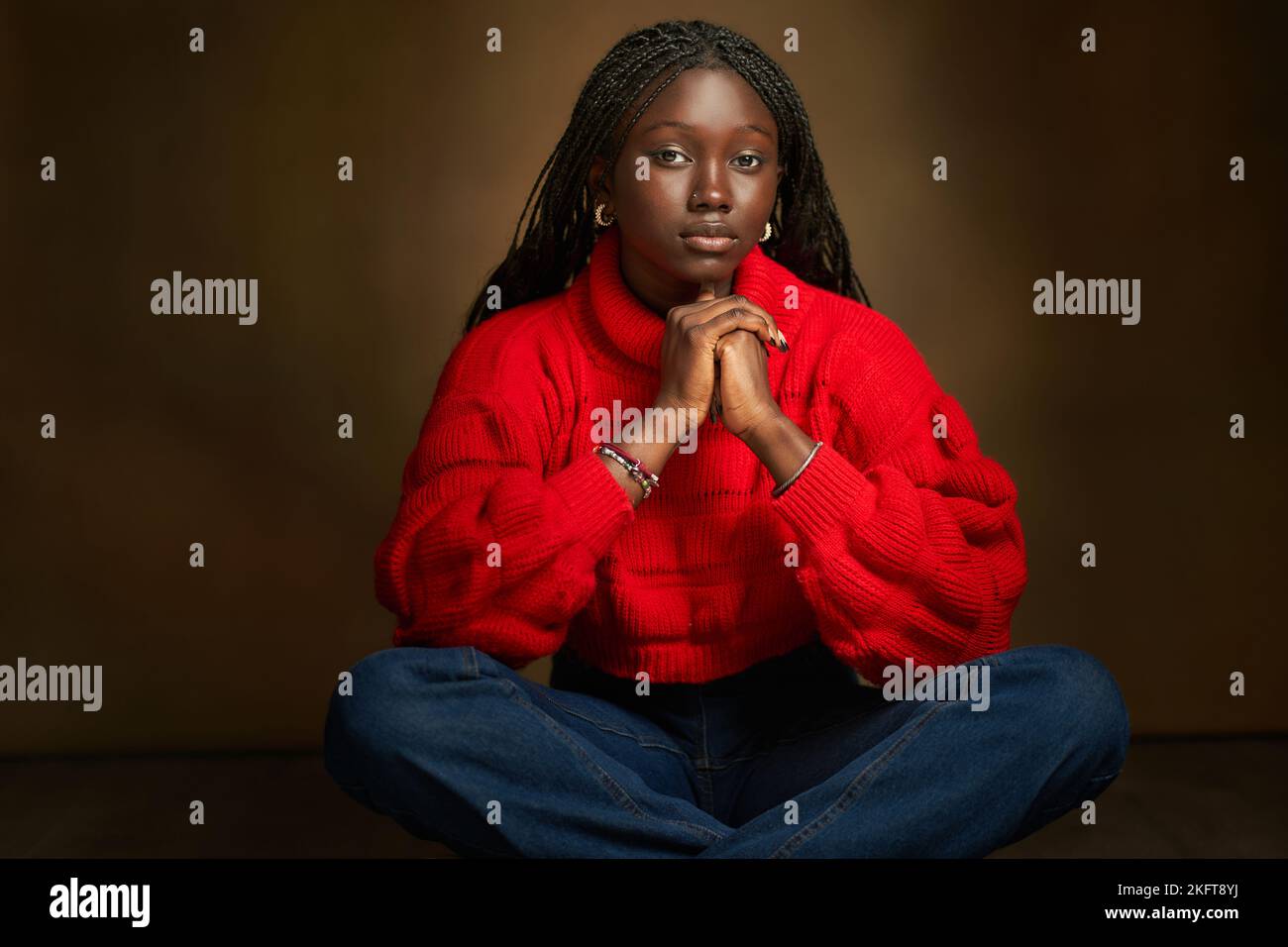 Pensive African American teenager sitting with legs crossed in studio on blurred brown background looking at camera thoughtfully Stock Photo