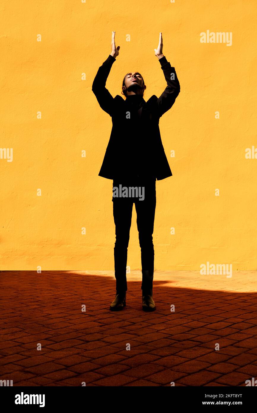 Young Hispanic male in black suit with long hair spreading arms and enjoying sunlight against vivid yellow wall Stock Photo