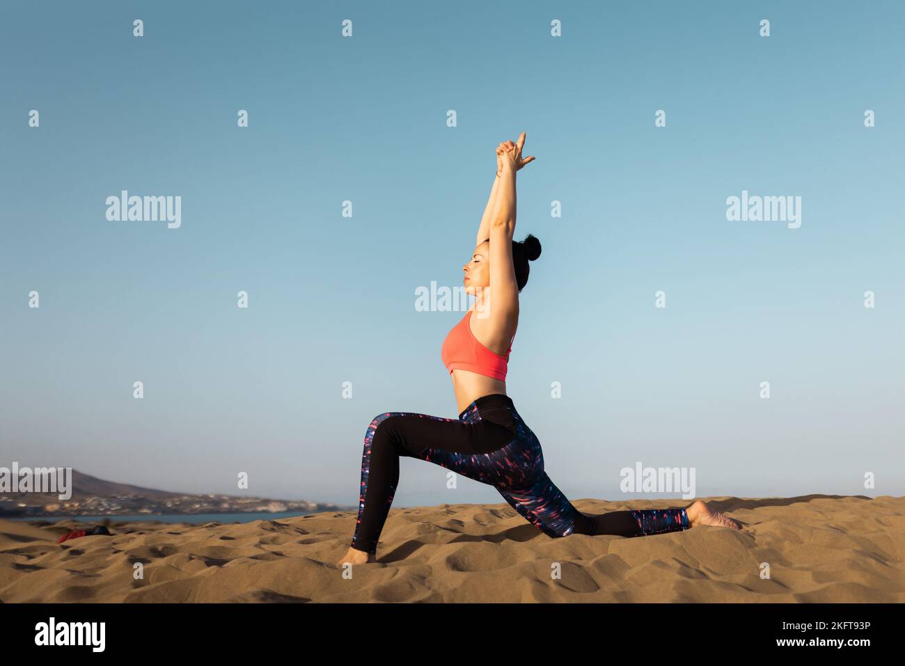 Side view of slim young woman in sportswear standing on sandy beach and stretching arms while practicing yoga on sunny day. Virabhadrasana posture Stock Photo