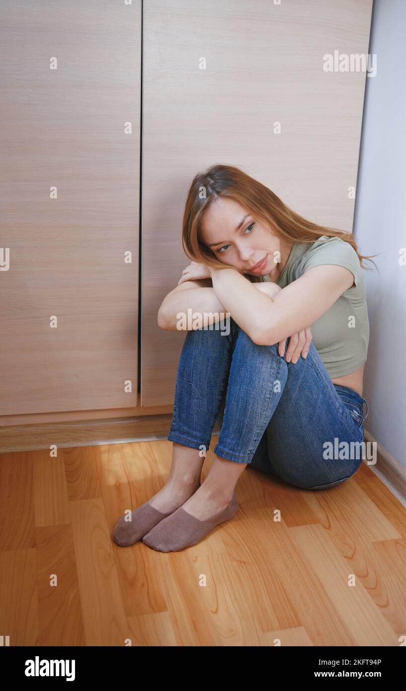 Full body high angle of thoughtful woman sitting on floor near wardrobe with arms crossed hugging knees looking away pensively Stock Photo