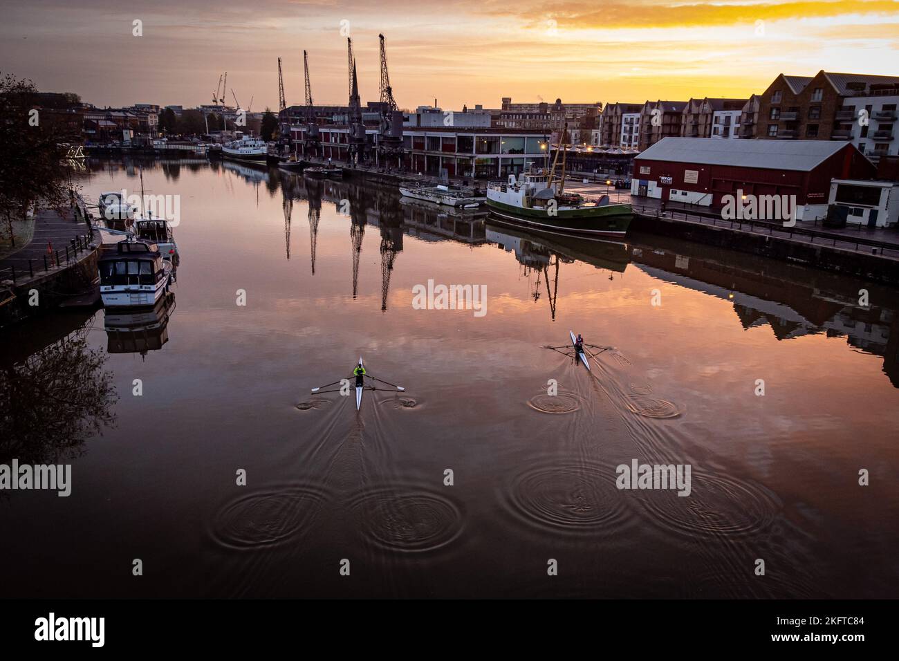 The sun rises over Bristol Harbourside as rowers leave a wake on the glassy water as they cross the basin past the old docks on a cold, but sunny morning across the south west. Picture date: Sunday November 20, 2022. Stock Photo