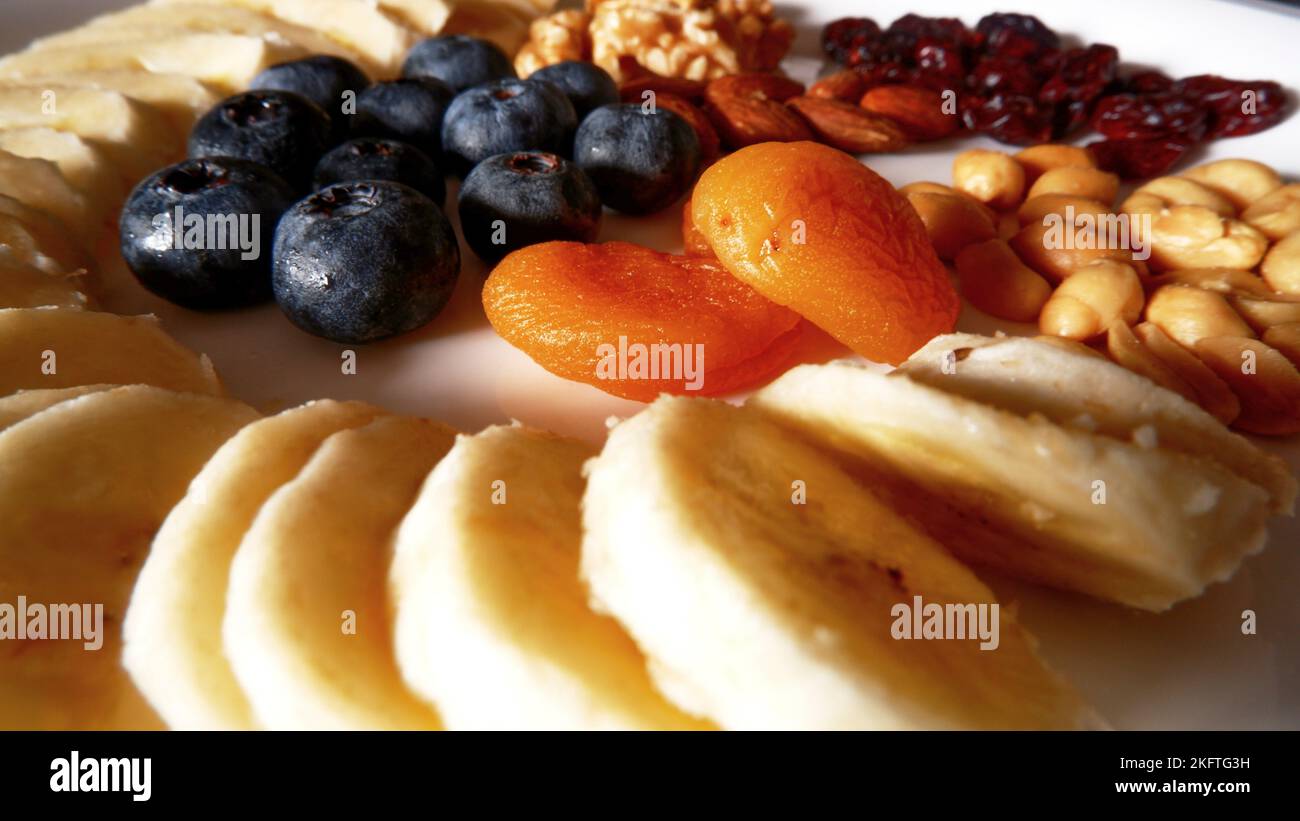 Fresh and dried fruits and nut dish, banana sliced, blueberries and apricot in the center and of the white plate with nut. Stock Photo