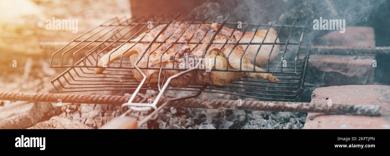 chicken meat food fried grill on smoldering coals or ember from campfire on ground on summer or autumn day. barbecue in camping conditions. survival o Stock Photo