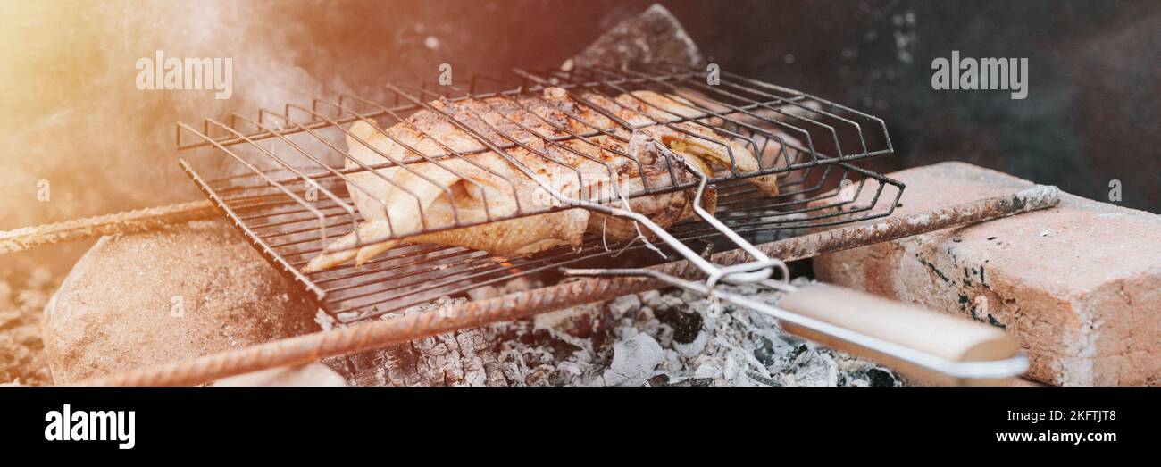 chicken meat food fried grill on smoldering coals or ember from campfire on ground on summer or autumn day. barbecue in camping conditions. survival o Stock Photo
