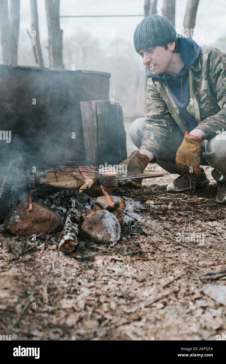 young man survivalist cooks roasts chicken meat food are fried on grill on smoldering coals or ember from a campfire on the ground. barbecue in campin Stock Photo