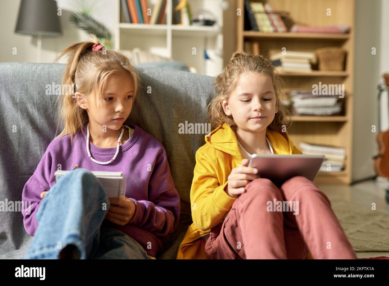 Lttle girl playing online on tablet pc sitting on floor together with her friend while she drawing in notebook Stock Photo