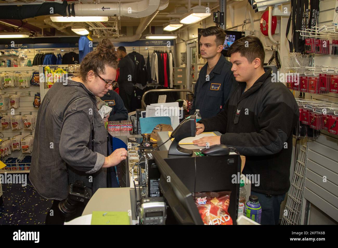 Retail Services Specialist 3rd Class Adriel Garcia-Castaneda, right, from Jonesboro, Georgia, and Retail Services Specialist Seaman Timothy Whiteside, from Valparaiso, Indiana, both assigned to the first-in-class aircraft carrier USS Gerald R. Ford’s (CVN 78) supply department, conduct a sale with Kendall Warner, a photographer with the Virginia-Pilot, during a visit to the ship store, Oct. 7, 2022. The Gerald R. Ford Carrier Strike Group (GRFCSG) is deployed in the Atlantic Ocean, conducting training and operations alongside NATO Allies and partners to enhance integration for future operation Stock Photo