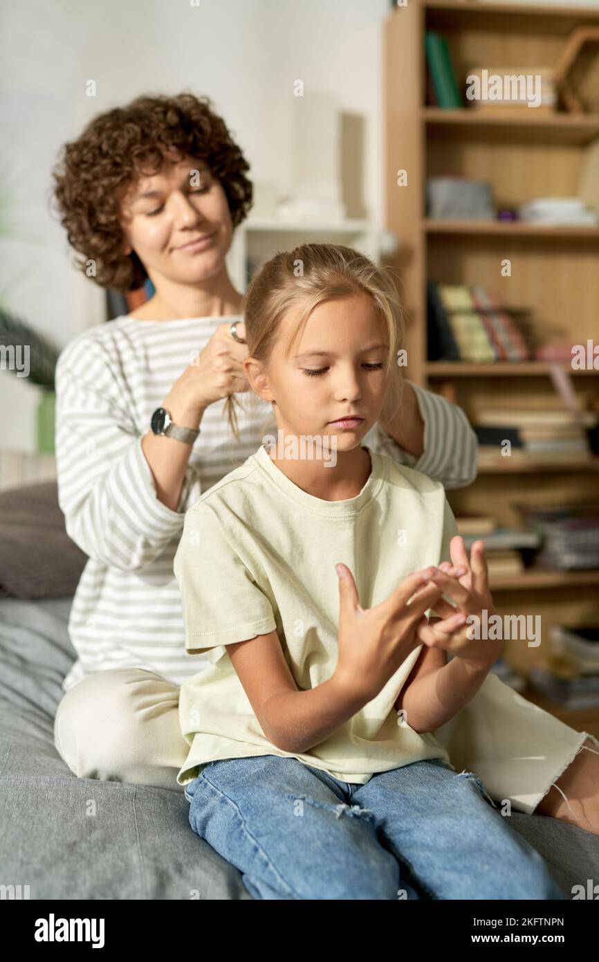 Young mother making hairstyle to her daughter while they sitting on bed