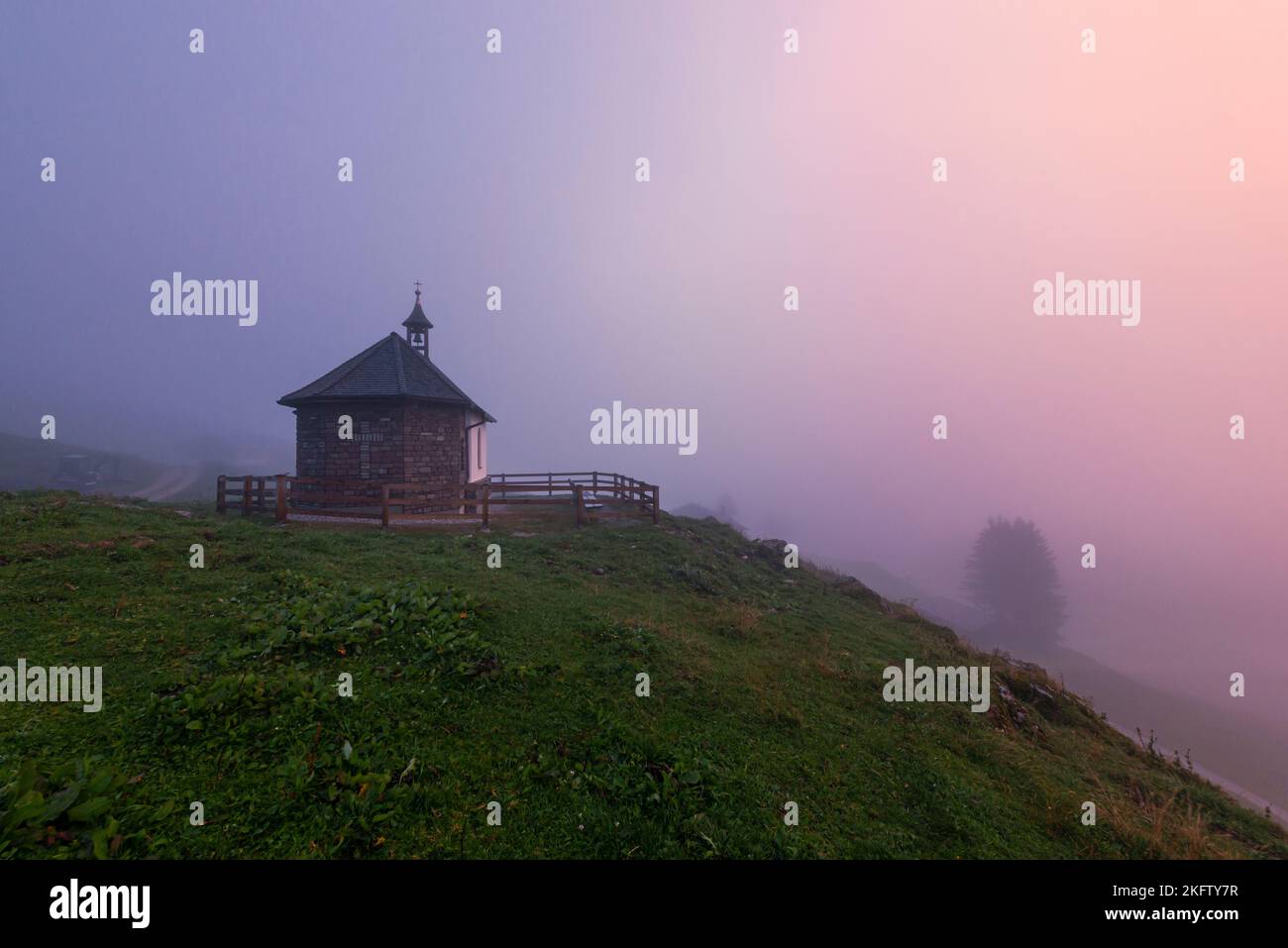 The reddish light of the sunset colors the thick foggy clouds at the chapel of the Ackernalm, Tyrol, Austria Stock Photo