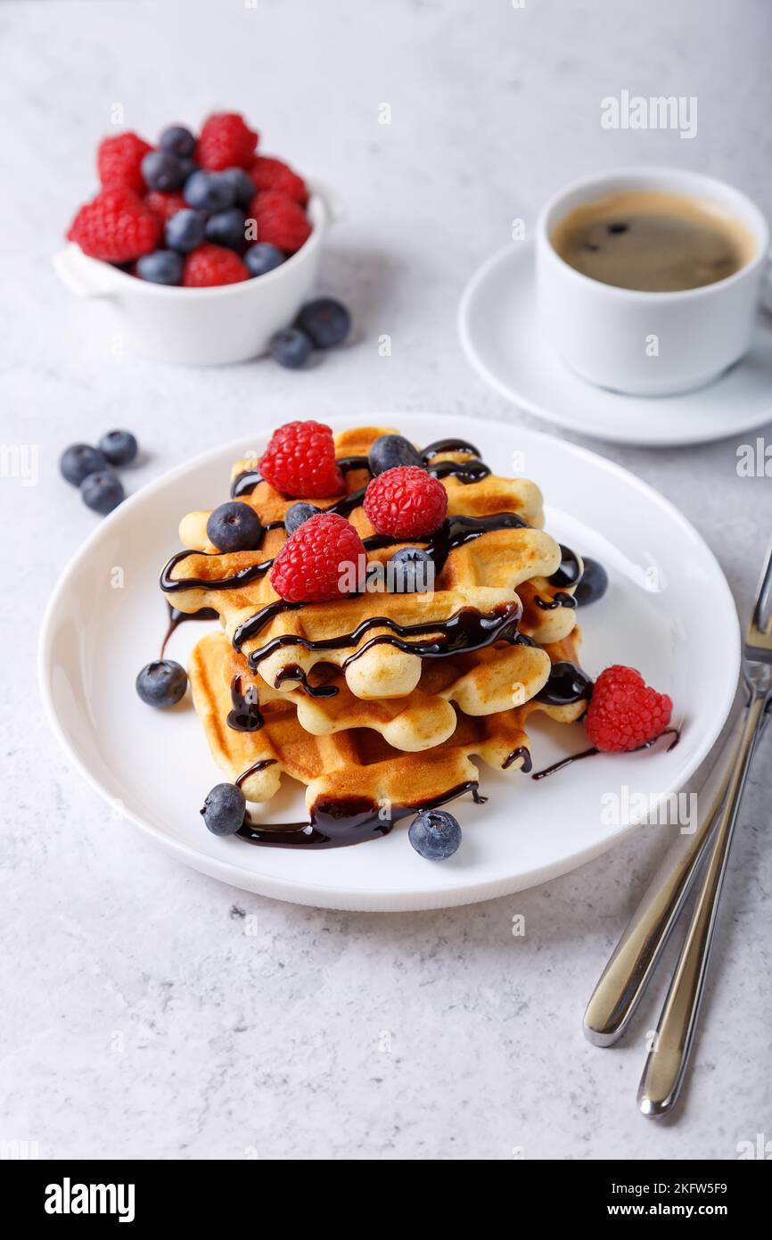 Viennese or Belgian waffles with fresh berries (raspberries and blueberries) and chocolate sauce on a white plate and a cup of coffee. Traditional des Stock Photo