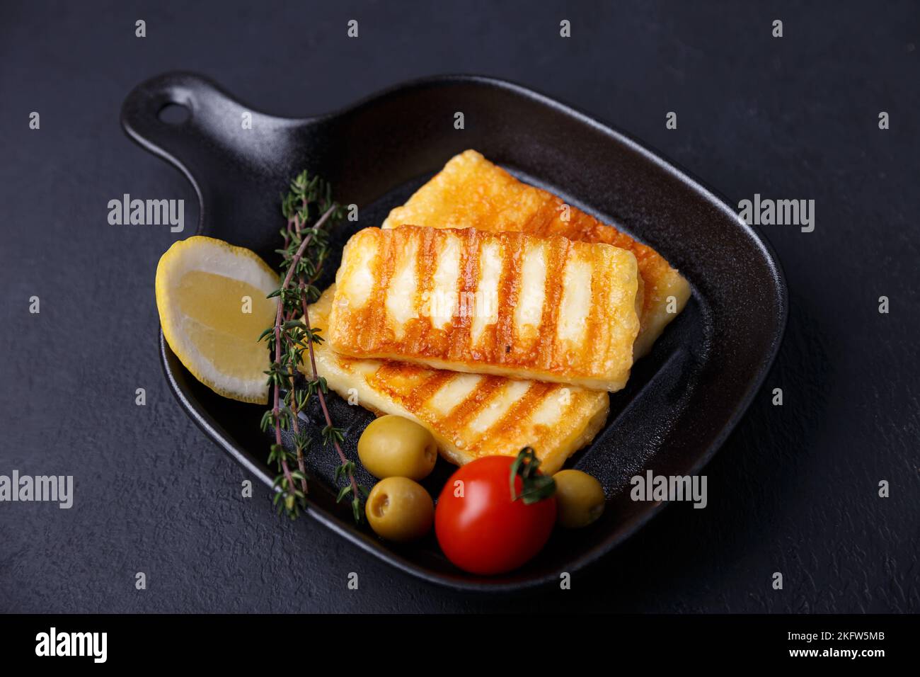 Grilled halloumi cheese in a black pan with olives, tomatoes, cucumbers, thyme and lemon