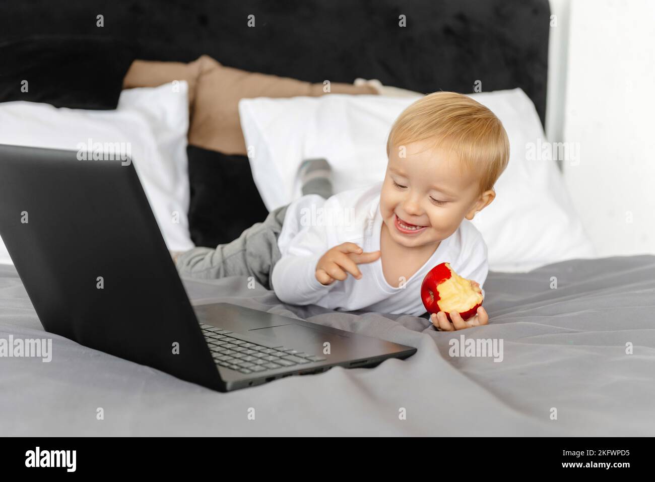 Cute kid is looking at a laptop and holding an apple, the rebonk is playing with the laptop Stock Photo