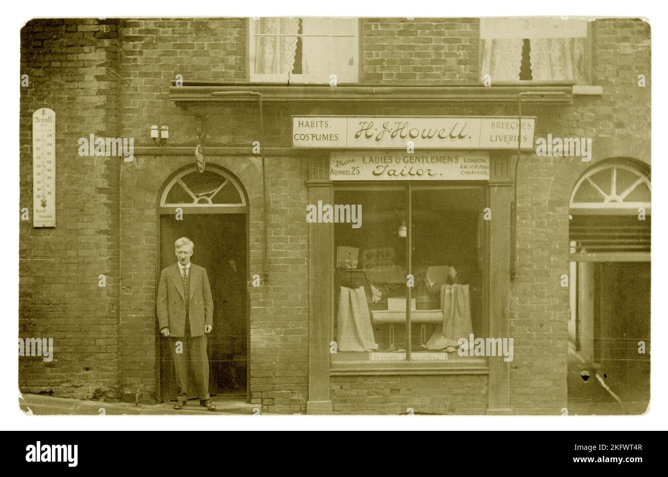Original WW1 era postcard of smartly dressed proprietor / tailor, standing in doorway outside a boutique shop - there's a sign for Burberry waterproofs in the window and lengths of tweed. Sign above states H J Howell Ladies and Gents tailors & also advertising tailoring of 'Costumes, Habits., Breeches & Liveries'  The sign also advertises other premises at 11 Milk St Cheapside London. But there is a telephone exchange on the sign for Arundel 25 - so this shop is in the West Sussex seaside town of Arundel in the High Street - perhaps he retired here and kept the London shop. Circa 1917, U.K. Stock Photo