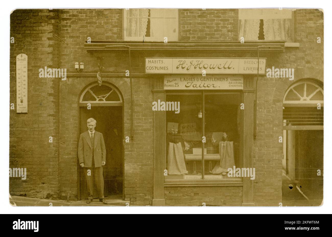 Original WW1 era postcard of smartly dressed proprietor / tailor, standing in doorway outside a boutique shop - there's a sign for Burberry waterproofs in the window and lengths of tweed. Sign above states H J Howell Ladies and Gents tailors & also advertising tailoring of 'Costumes, Habits., Breeches & Liveries'  The sign also advertises other premises at 11 Milk St Cheapside London. But there is a telephone exchange on the sign for Arundel 25 - so this shop is in the West Sussex seaside town of Arundel in the High Street - perhaps he retired here and kept the London shop. Circa 1917, U.K. Stock Photo