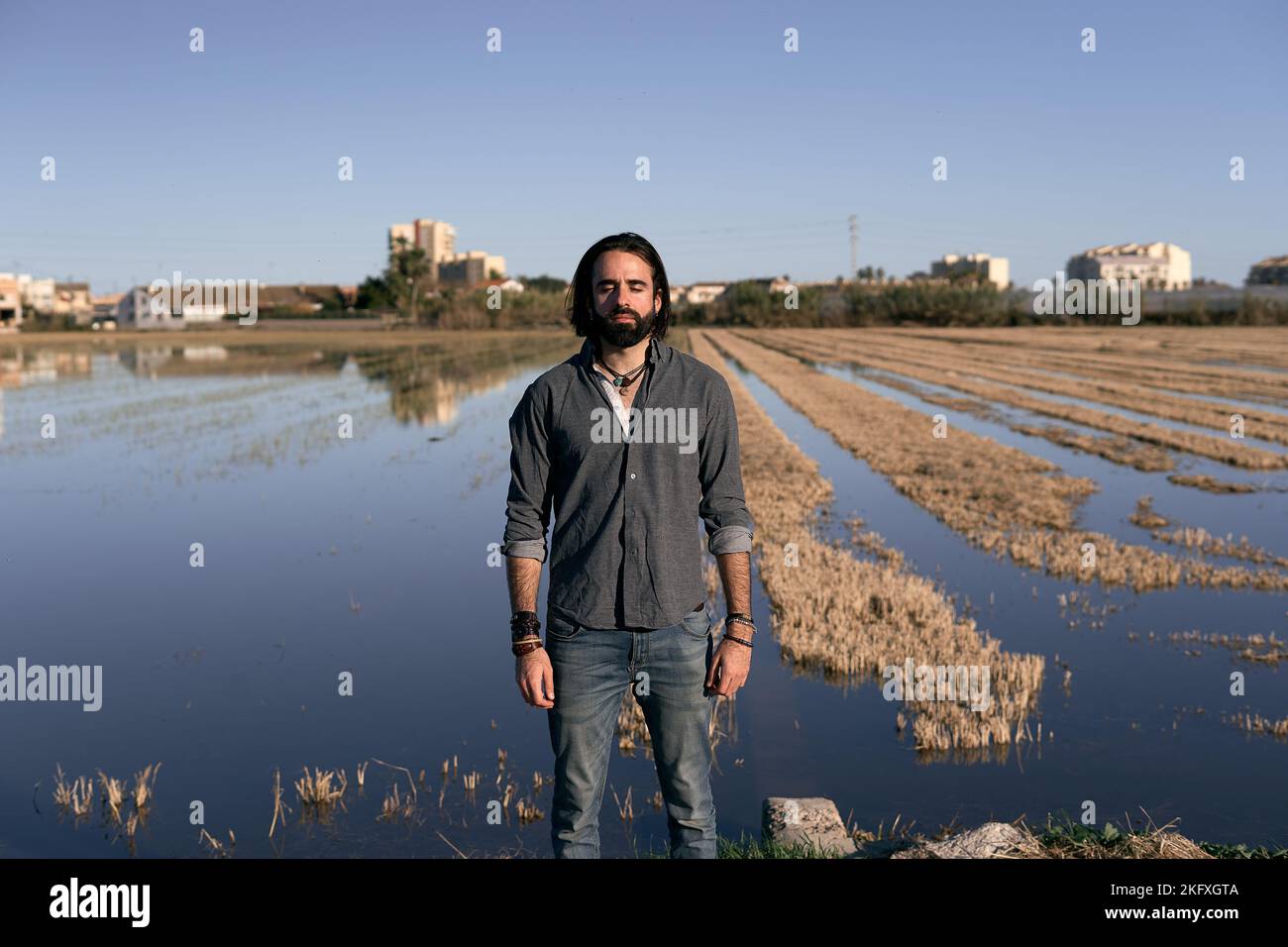 young caucasian man with long hair beard and gray shirt looking at camera with closed eyes in a field flooded with water Stock Photo