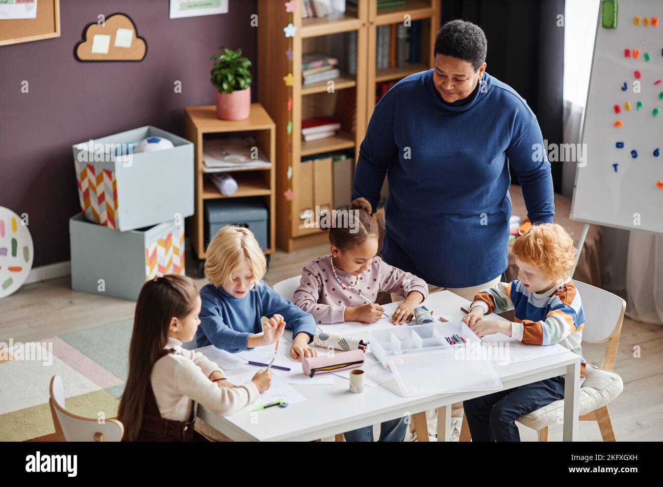 Group of adorable learners of nursery school sitting by table at lesson while mature African American female teacher standing by them Stock Photo