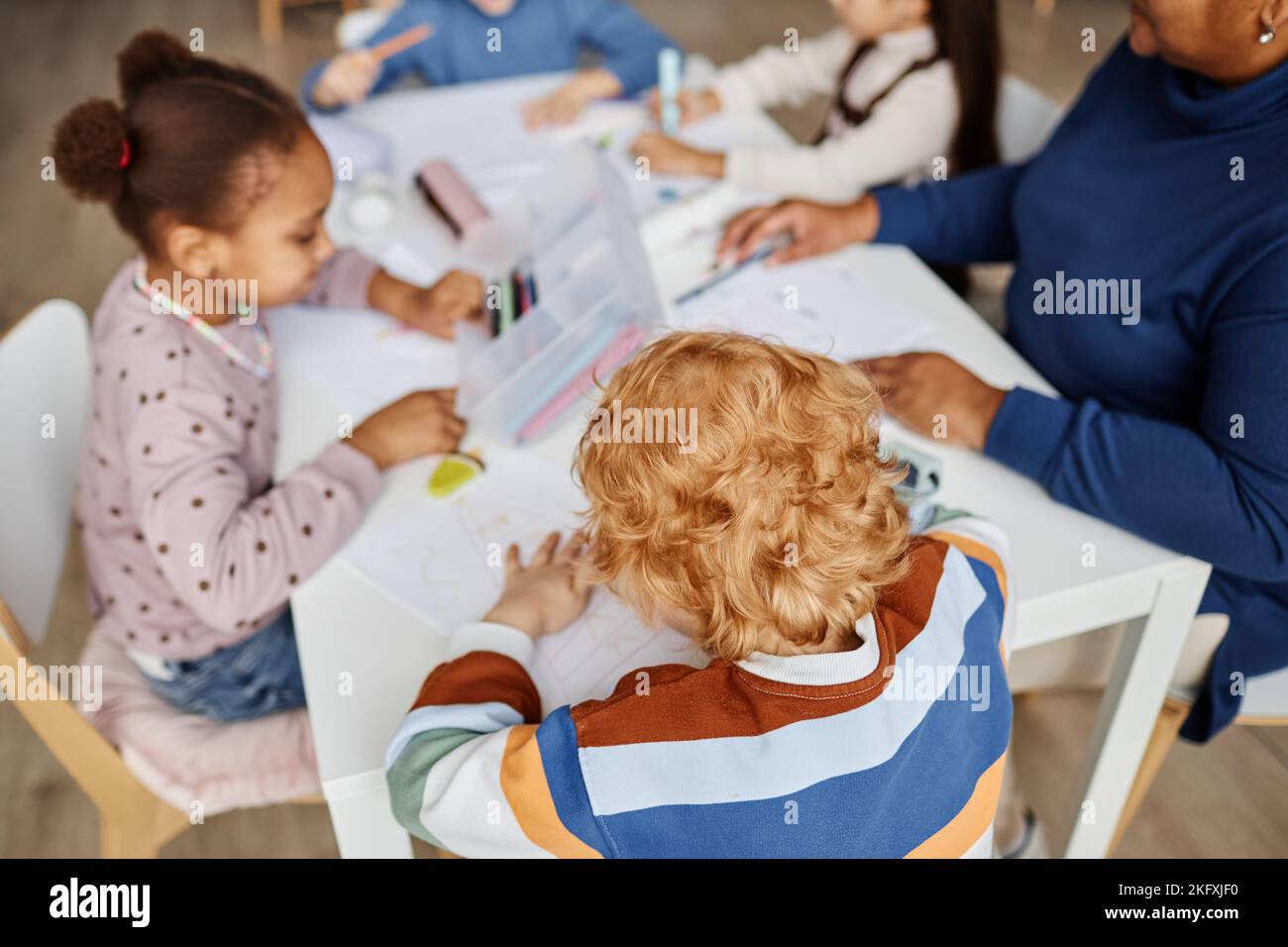 Above angle of diligent ginger haired boy drawing among intercultural classmates and teacher sitting by table at lesson in primary school Stock Photo
