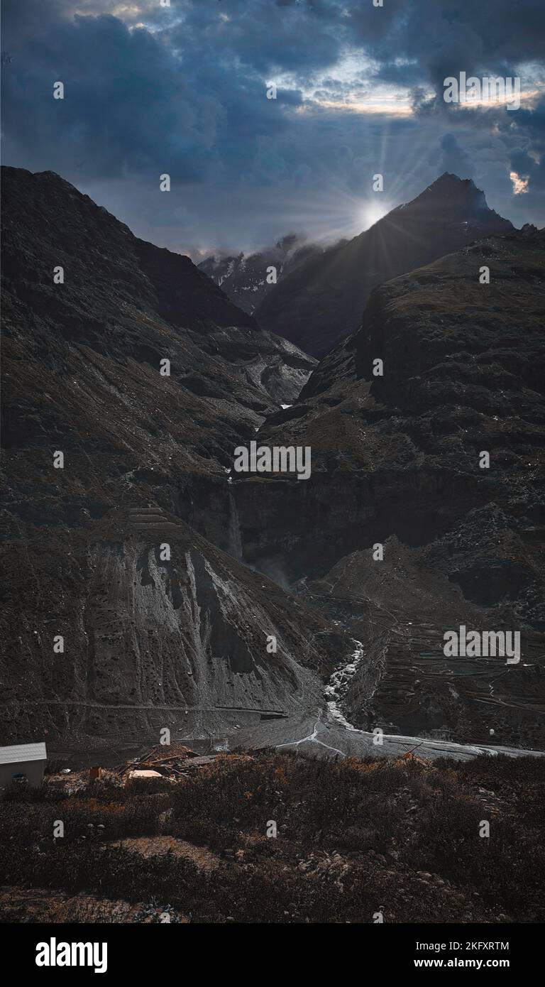 A vertical panoramic of the sun rising behind tall mountains of India and a small river coming through them Stock Photo