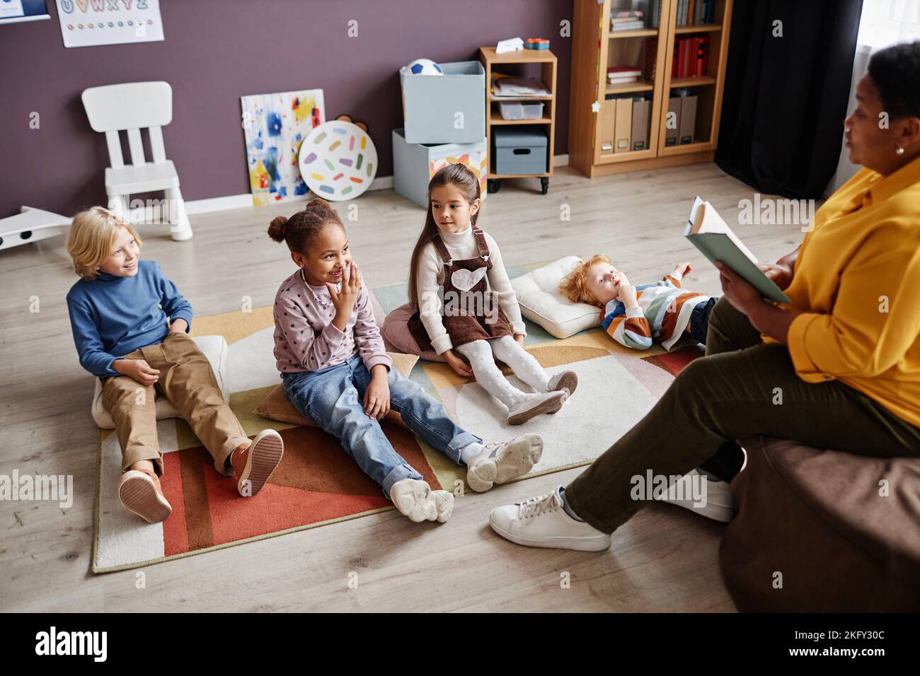 Group of curious intercultural kids relaxing on carpet in front of teacher reading them fairy tale in the miidle of the day at nursery school Stock Photo