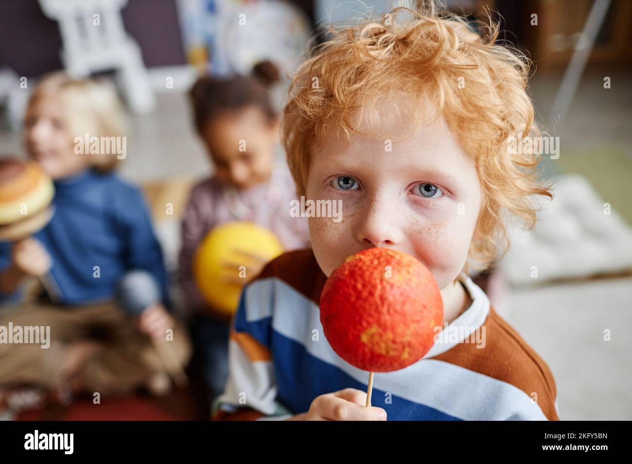 Adorable boy with ginger hair holding model of Mars planet and looking at camera while playing against other learners of nursery school Stock Photo