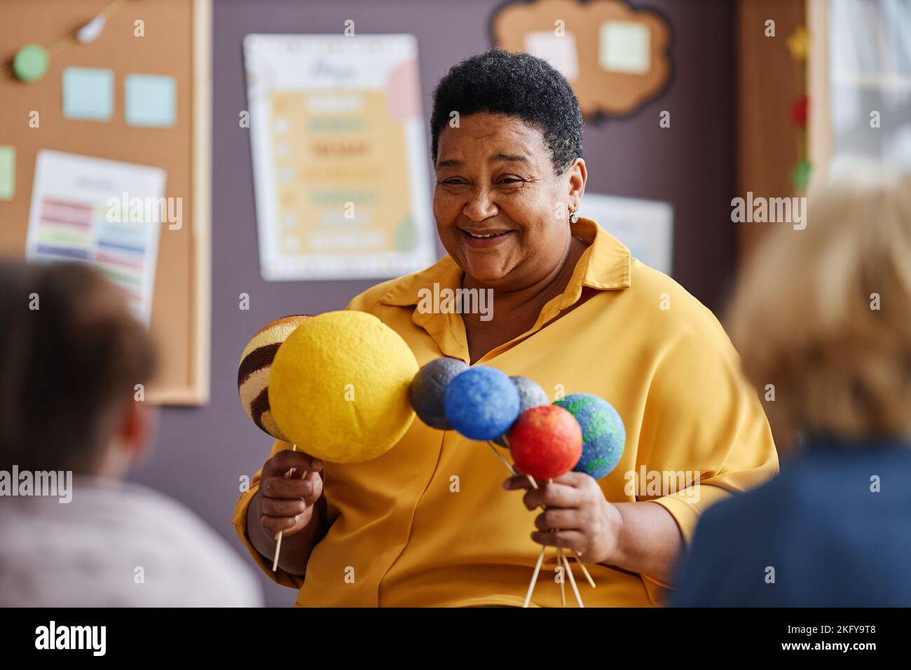 Happy mature African American teacher with planet models speaking about solar system to little learners of nursery school Stock Photo