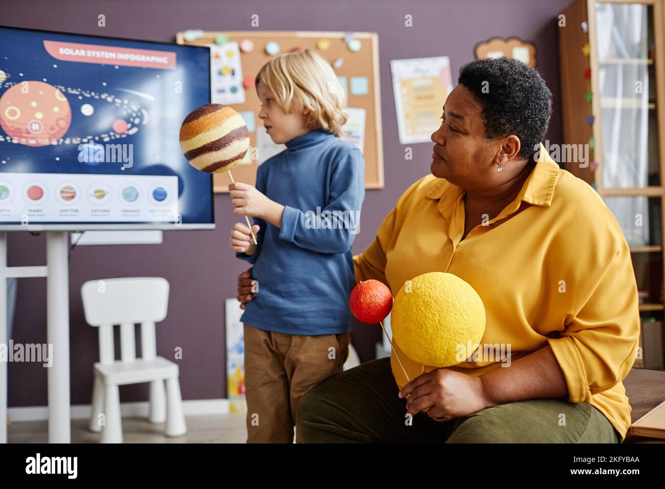 Mature teacher of nursery school and little schoolboy with planet models looking at whiteboard with poster representing solar system Stock Photo