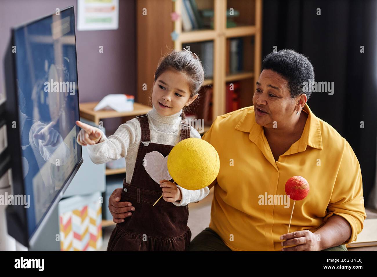 Clever little girl with model of Sun pointing at interactive board during presentation of solar system and discussion with teacher Stock Photo