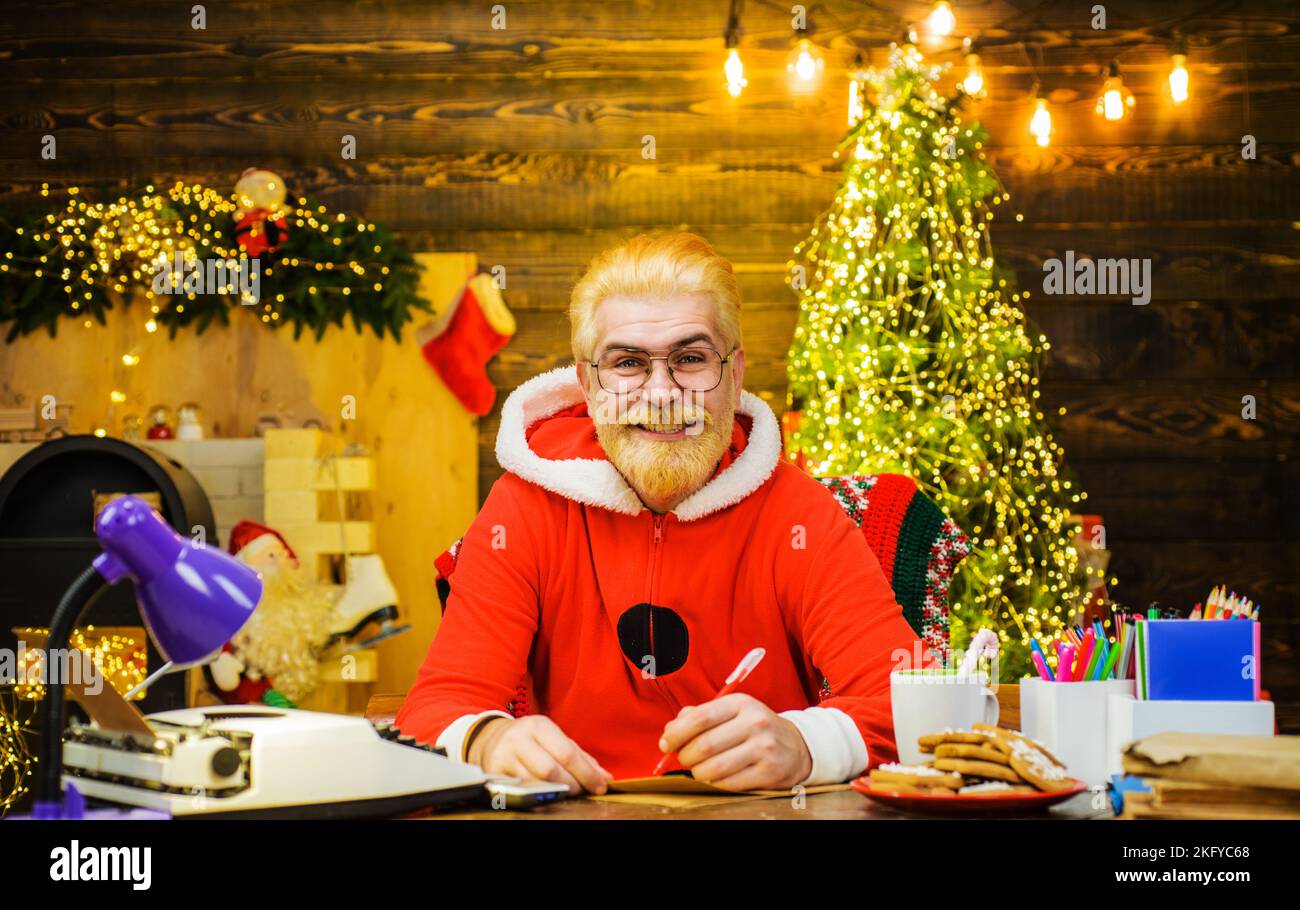 Happy bearded Santa Claus writing on wish list sitting home at workshop table. Christmas. New year. Stock Photo
