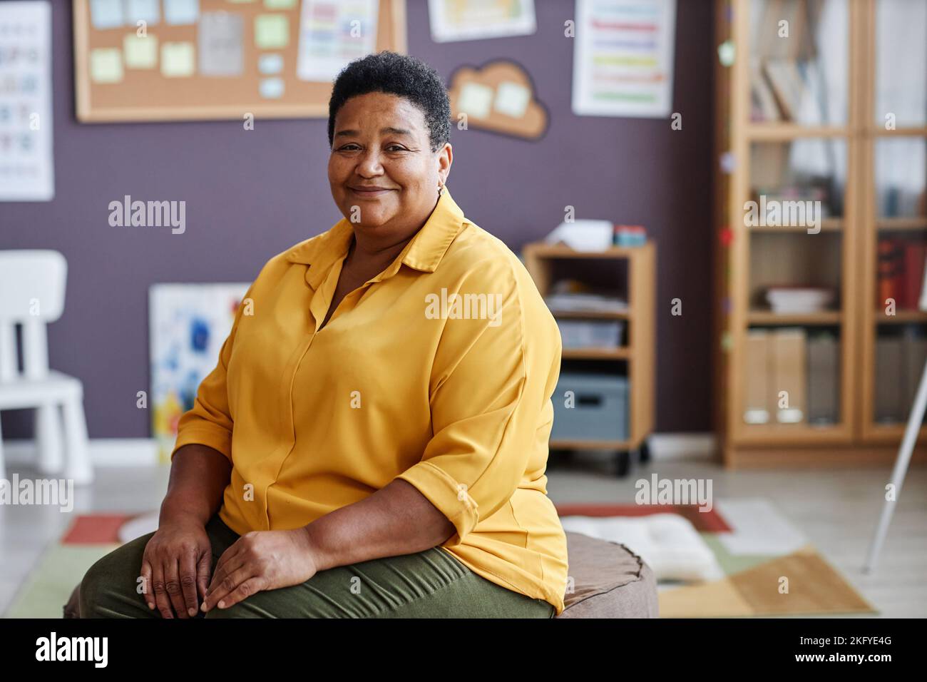 Portrait of experienced African American female teacher or coach in yellow shirt sitting in front of camera in classroom of nursery school Stock Photo