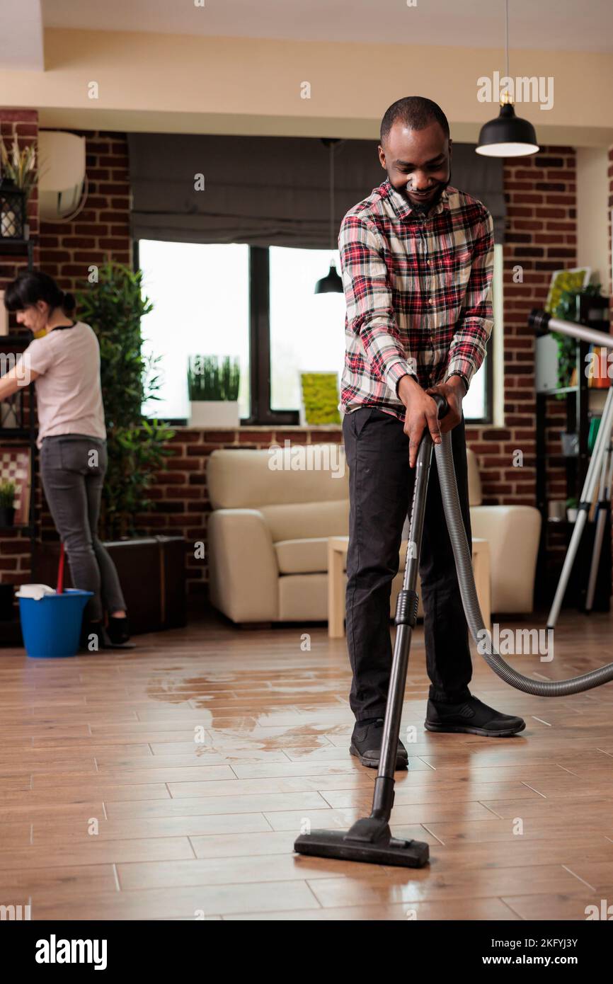 Smiling African American man vacuuming the floor while woman dusts the shelves. Multiracial couple cleaning the house on a saturday morning, doing the weekly cleaning routine. Household chores. Stock Photo
