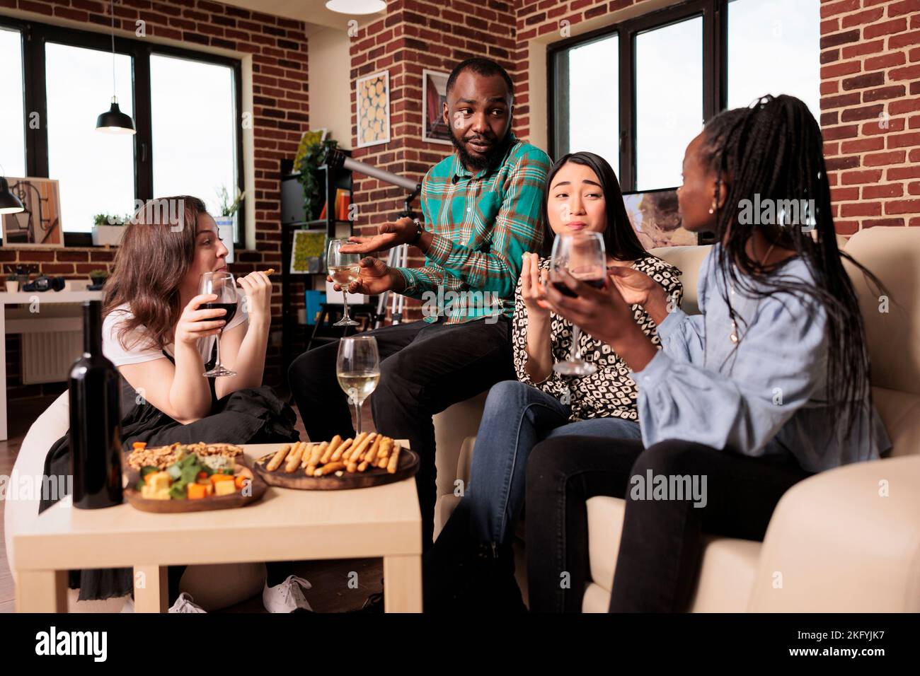 Culturally diverse friends talking, conversing each other, drinking wine, eating, tasting cheese, bread sticks at diverse people gathering. Different nationalities mates enjoying social life. Stock Photo