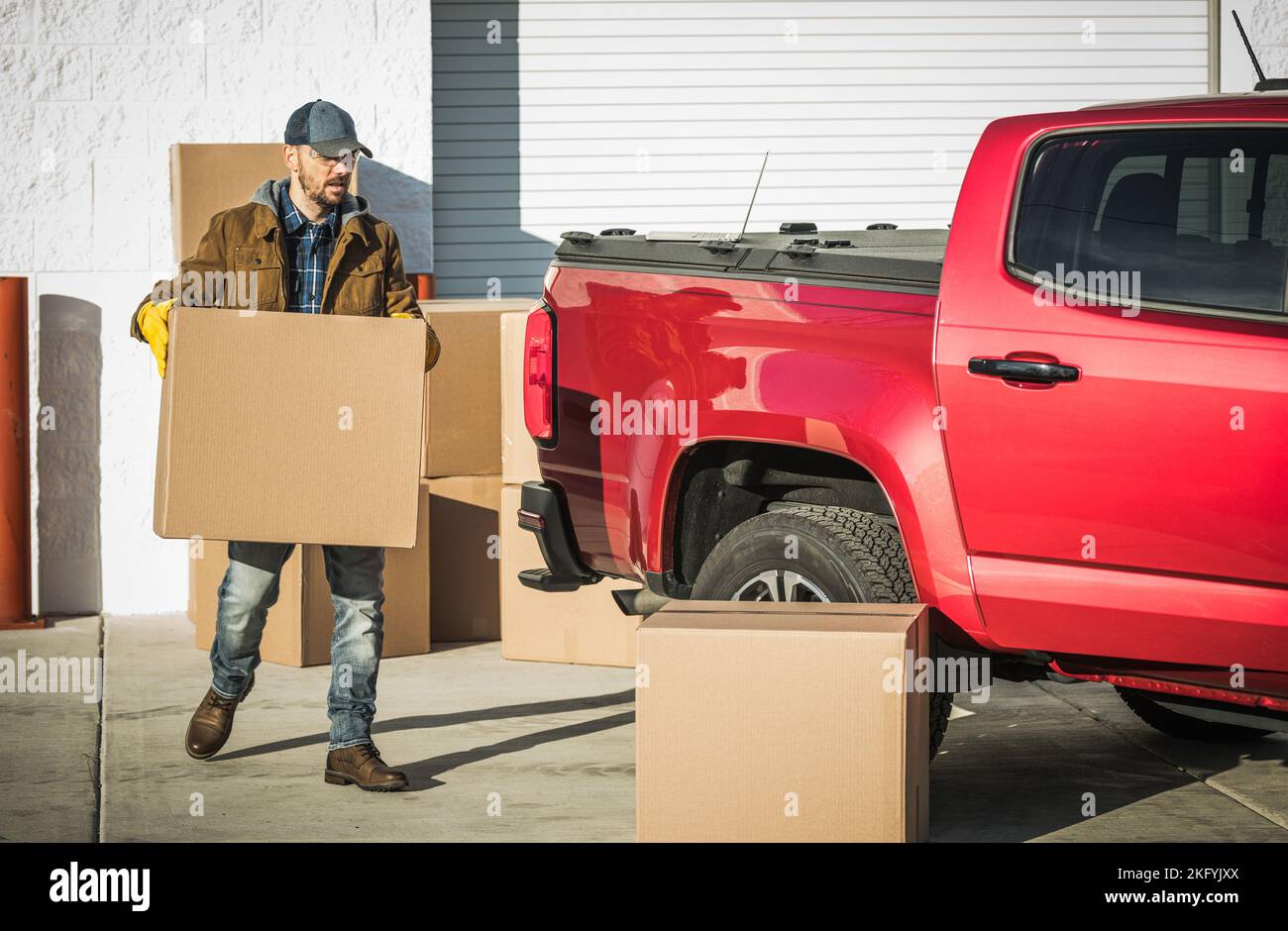 Professional Middle Aged Moving Company Worker Carrying a Cardboard Box with Client's Belongings to His Big Red Pickup Truck. Relocation Services Them Stock Photo