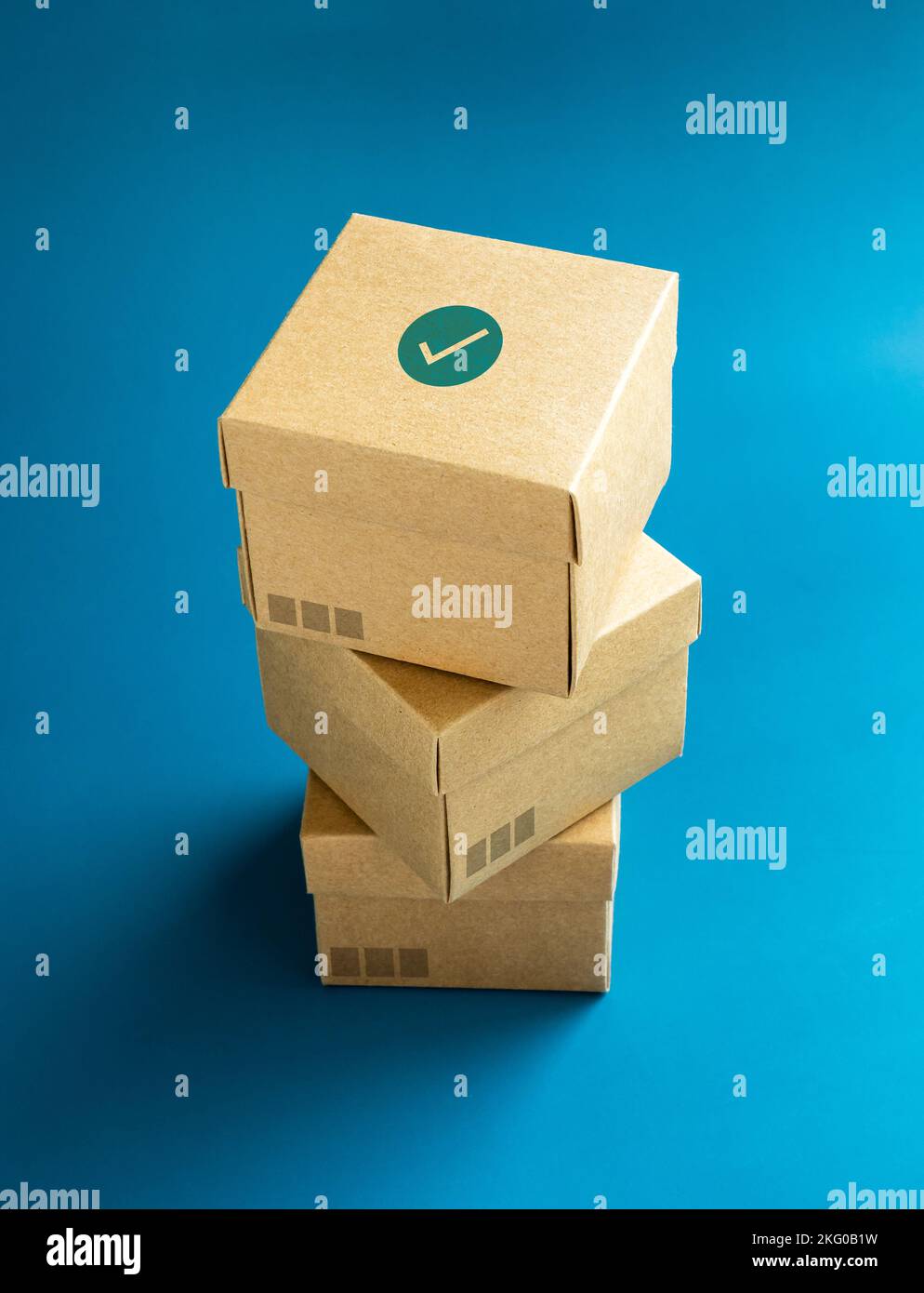 Green check mark icon on lid of cardboard parcel boxes stack on blue background, vertical style. Carton kraft paper box with lid on top. Delivery, pac Stock Photo