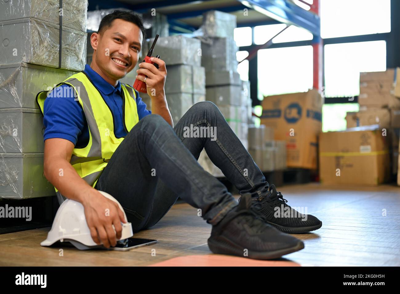 Handsome and happy millennial Asian male warehouse worker in uniform sitting in warehouse, using
