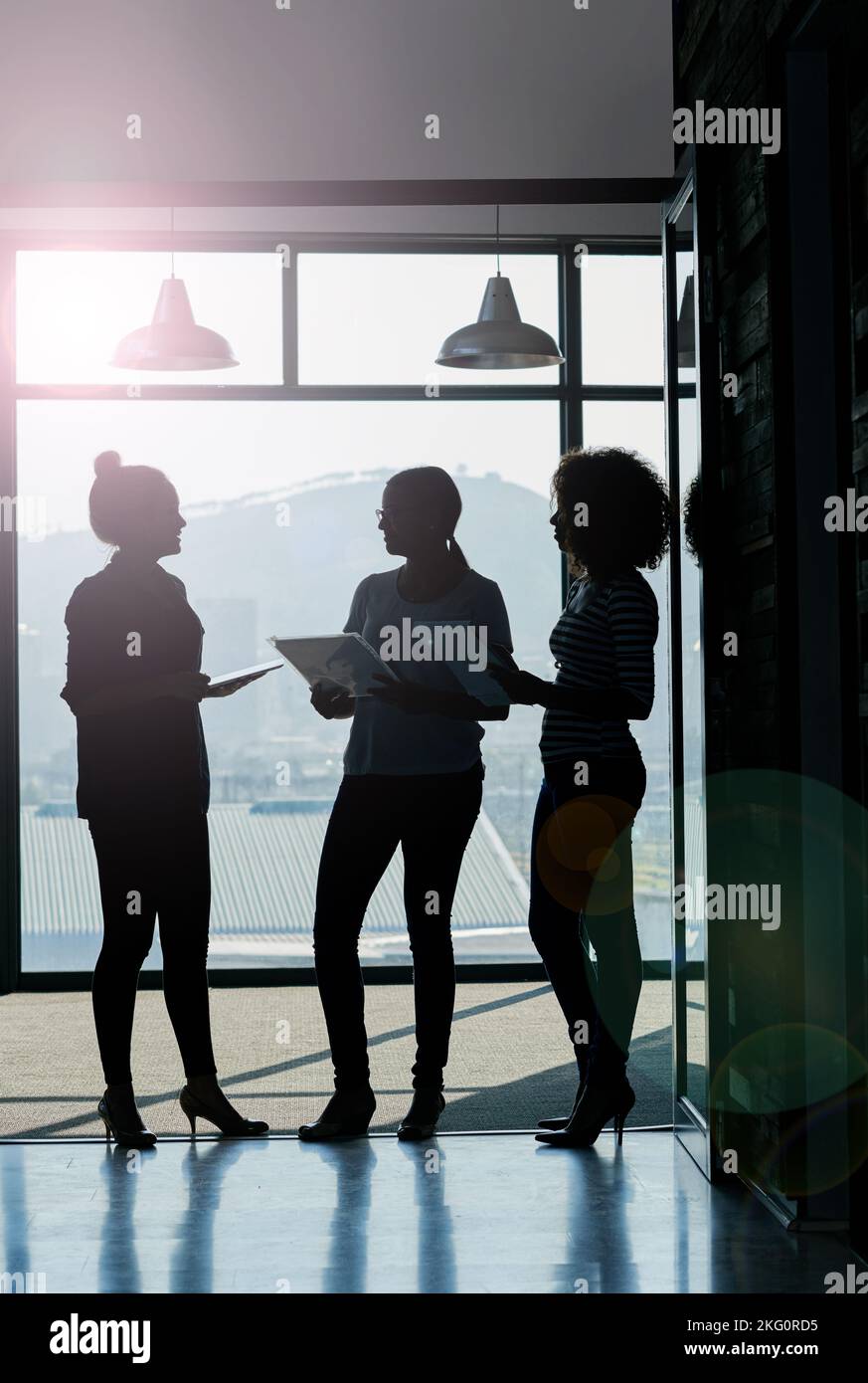 Think well be working over time on this...Silhouette shot of female coworkers talking while standing in front of a window in an office. Stock Photo