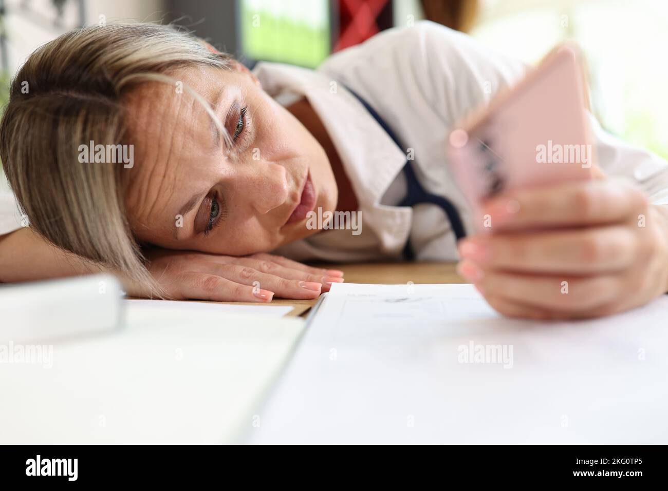 Frustrated female doctor putting head on table and surfing internet on smartphone Stock Photo