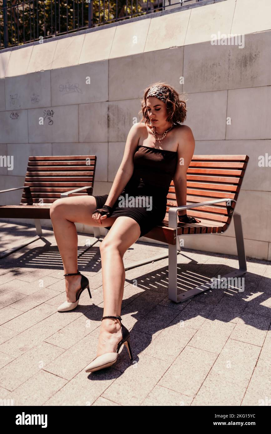 Portrait of a woman on the street. An attractive woman in a black dress is sitting on a bench outside. Stock Photo