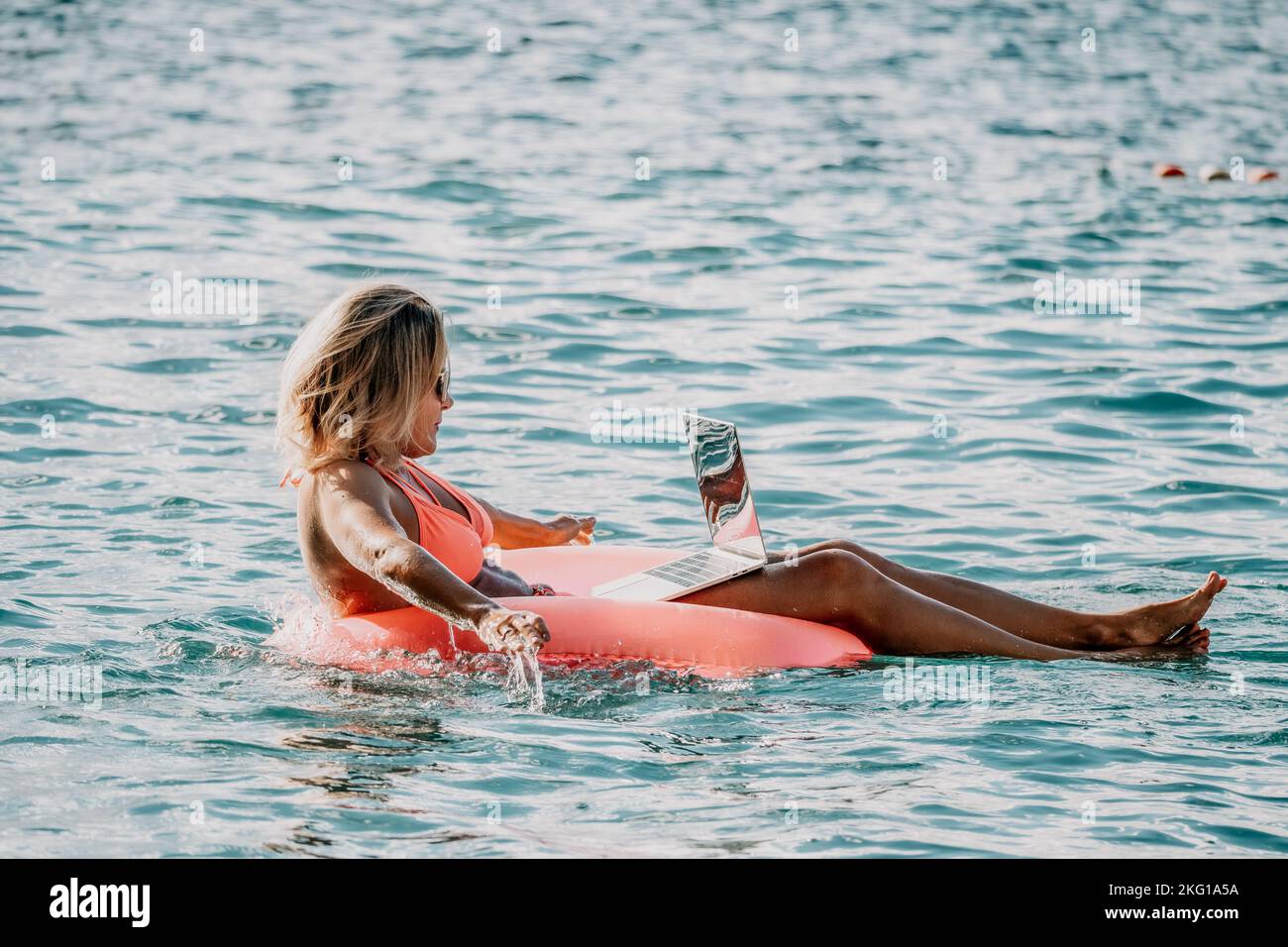 Woman works on laptop in sea. Freelancer, young blond woman in sunglases floating on an inflatable big pink donut with a laptop in the sea at sunset Stock Photo