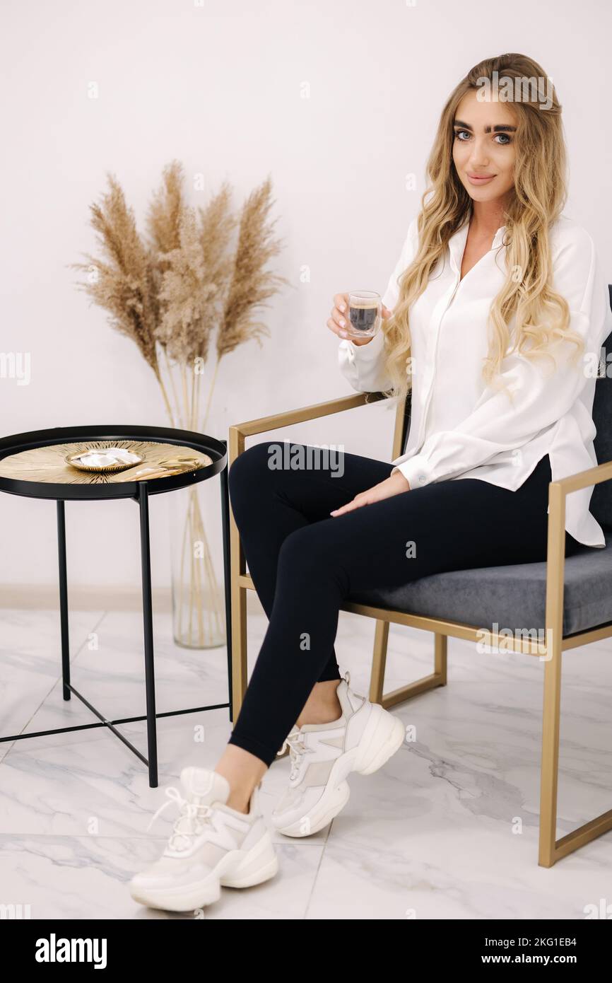 Gorgeous model waiting for cosmetology procedure, drink coffee and use phone. White interior of beauty studio. Blond hair woman Stock Photo