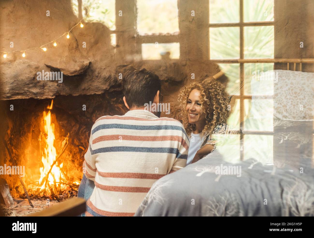 One couple man and woman enjoy leisure indoor activity sitting in front of a warm fireplace to heat home. Happy young mature people view through a gla Stock Photo