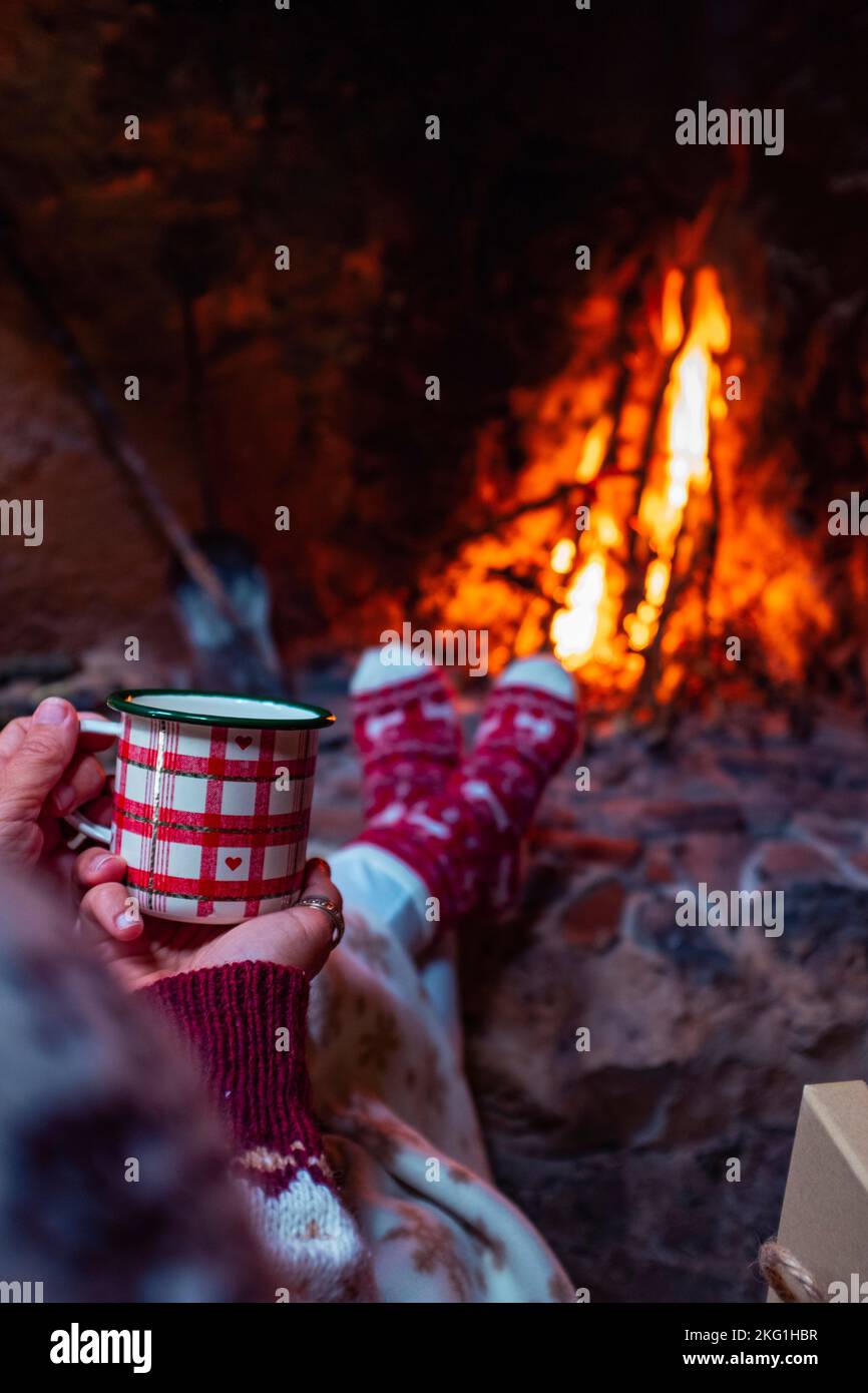 Close up of people holding christmas mug cup and drinking in front of a warm fireplace at home in chalet for december holiday season vacation. Female Stock Photo