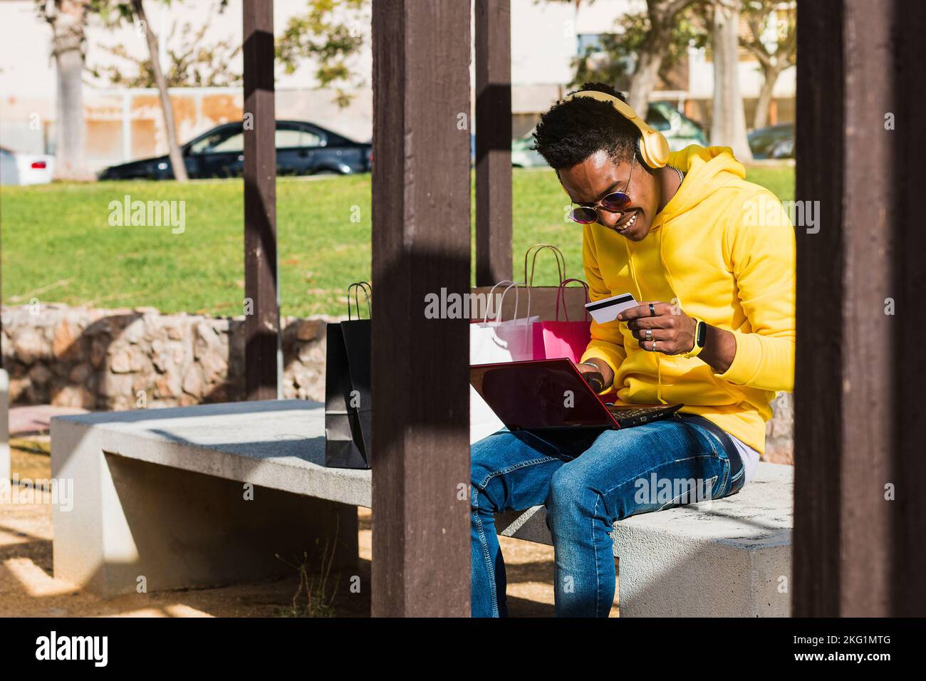 A smiling young African man sits on a park bench next to some shopping ...