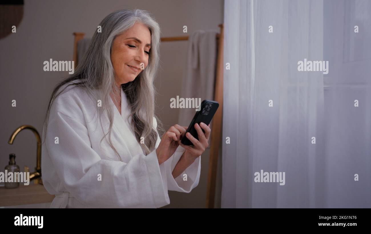 60s old Caucasian senior mature woman grandmother lady standing in bathroom in bathrobe looking at phone booking ordering cosmetics cream for Stock Photo
